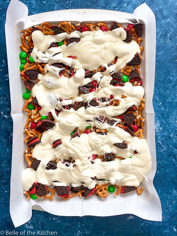 blue background with a sheet pan covered in white paper, full of M&Ms, Oreos, and pretzels with melted almond bark drizzled on top