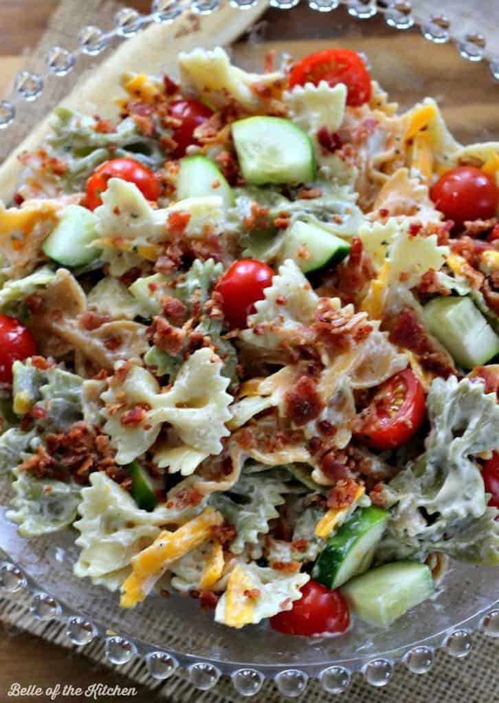 A close up of a bowl of pasta salad topped with cucumber, bacon, tomatoes, and shredded cheese