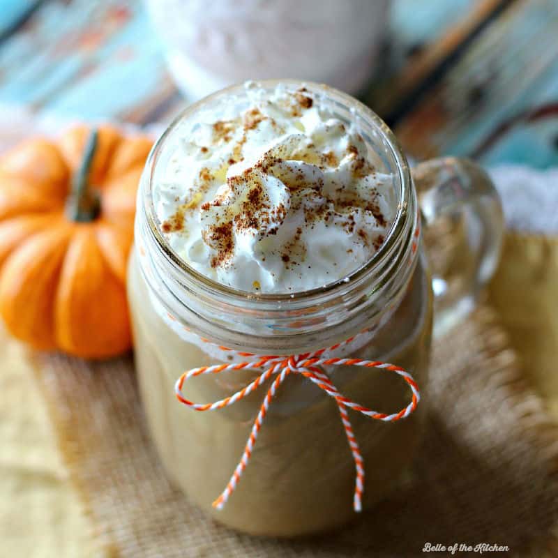 A mug filled with coffee and whipped cream wrapped in an orange piece of string with a pumpkin in the background