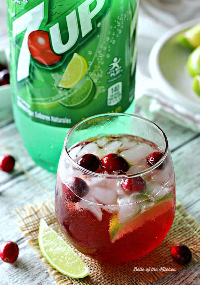 A glass of limeade with cranberries and a lime slice and a bottle of 7up in the background