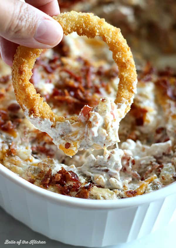 A dish filled with cheeseburger dip topped with bacon and an onion ring dipped into it