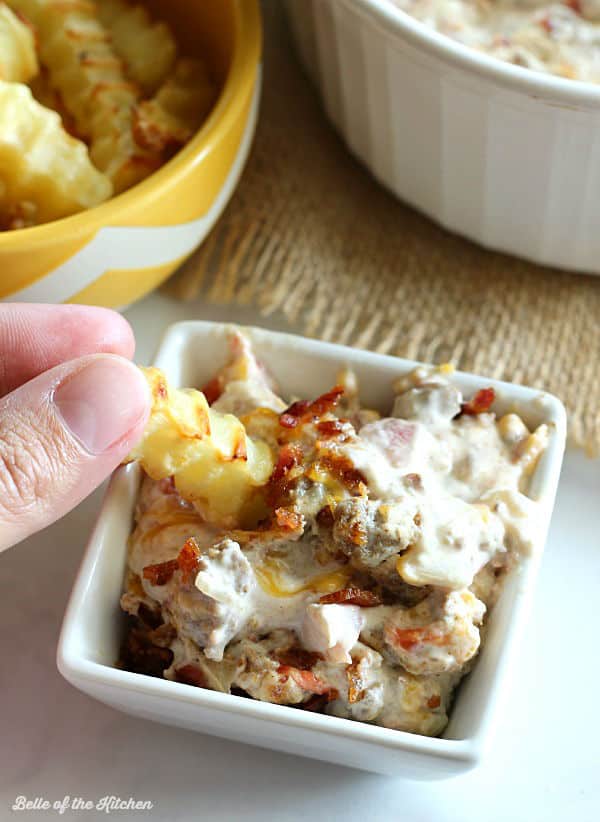 A dish filled with cheeseburger dip topped with bacon with a fry dipped in