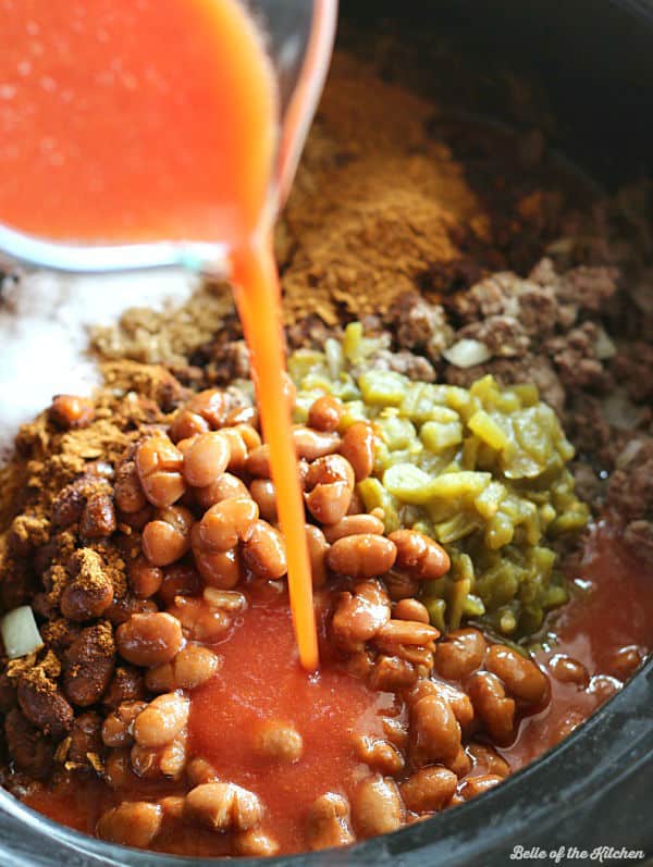 A crockpot full of beans, green chiles, ground beef, and more with tomato juice being poured in