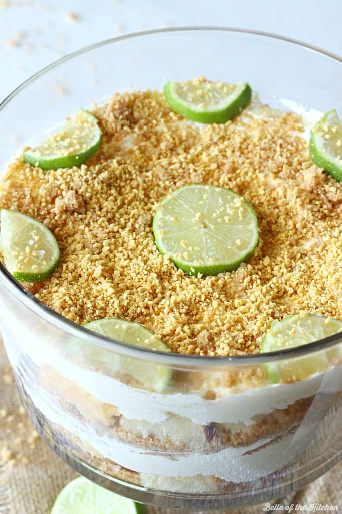 A close up of a trifle dish filled with layers of cheesecake filling, chopped pound cake, and topped with graham cracker crumbs and sliced lime