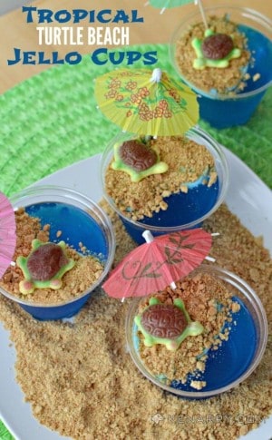 A plate of blue jello cups topped with graham crackers, candy turtles, and drink umbrellas