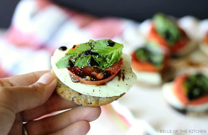 a hand holding a cracker topped with sliced tomato, mozzarella, basil, and balsamic glaze