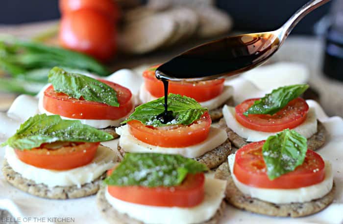 a spoon pouring balsamic glaze over crackers topped with sliced tomato, mozzarella, and basil