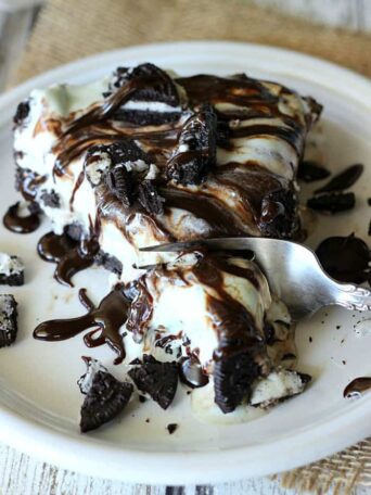 A piece of chocolate ice cream cake on a plate, with Chocolate fudge and Oreos