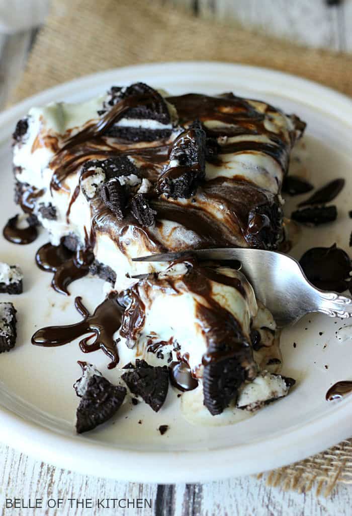 A piece of chocolate ice cream cake on a plate, with Chocolate fudge and Oreos 