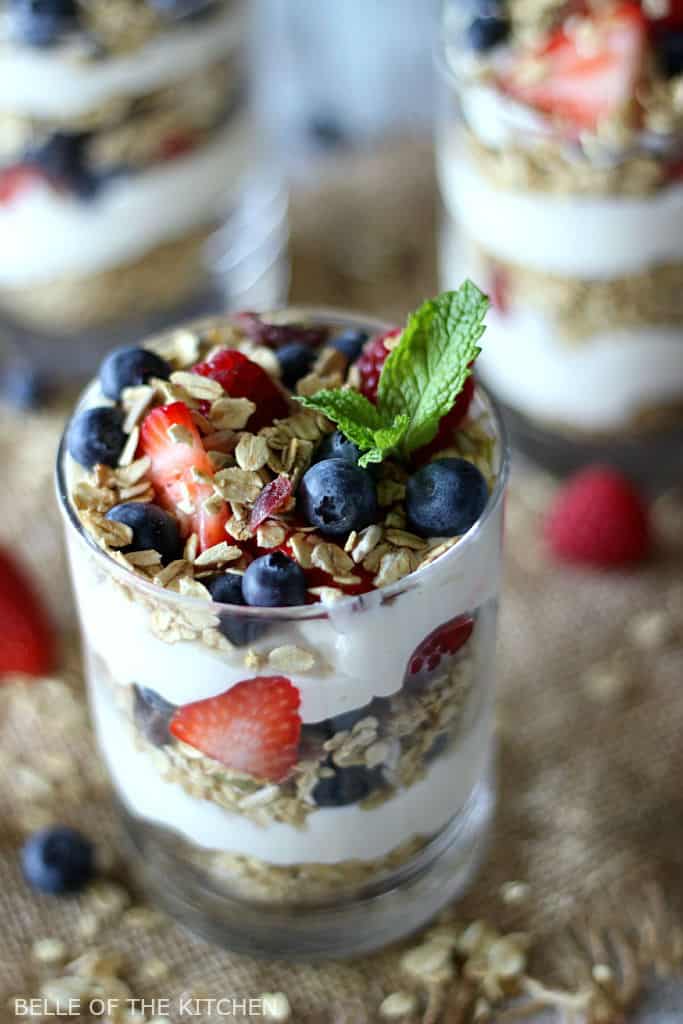 A glass filled with layers of cheesecake, fruit, and granola, topped with mint and a spoon