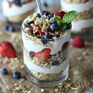 A glass filled with layers of cheesecake, fruit, and granola, topped with mint and a spoon