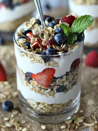 A glass filled with layers of cheesecake, fruit, and granola, topped with mint and a spoon