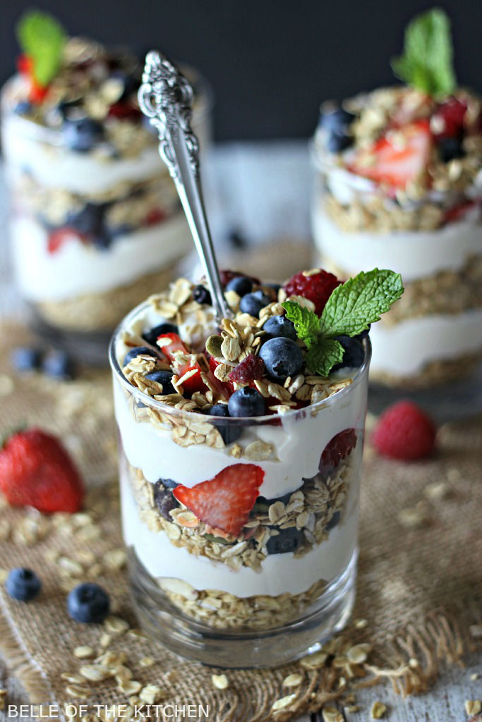 A glass filled with layers of cheesecake, fruit, and granola, topped with mint and a spoon