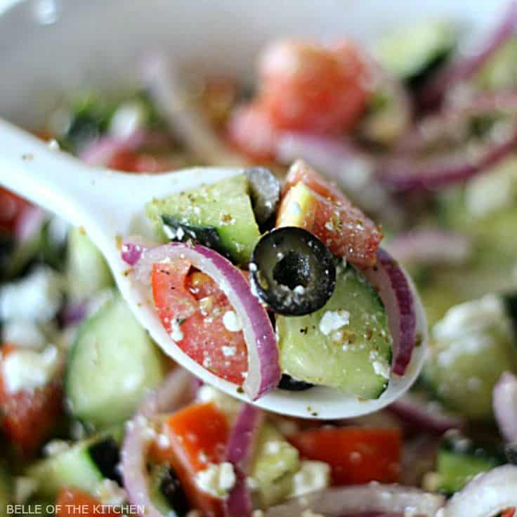 A close up of a spoon filled with tomatoes, cucumbers, red onion, black olives, and feta cheese
