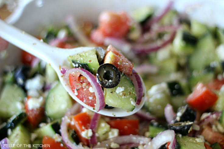 A close up of a spoon filled with tomatoes, cucumbers, red onion, black olives, and feta cheese