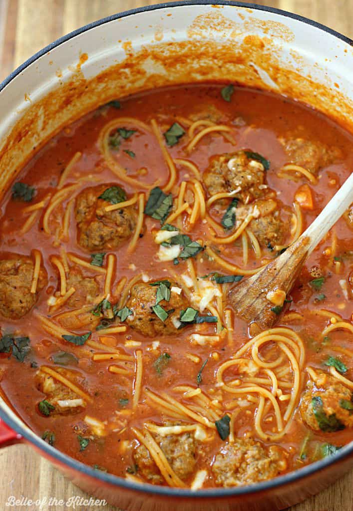 A pot filled with soup, meatballs, and spaghetti noodles topped with cheese and basil