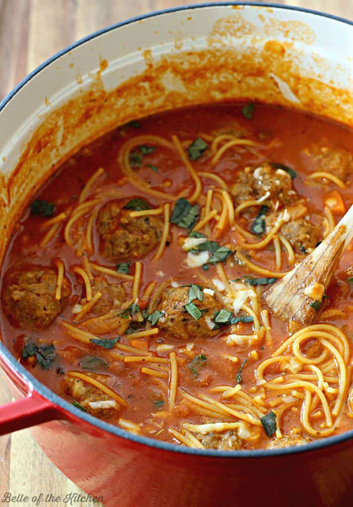 A pot filled with soup, meatballs, and spaghetti noodles topped with cheese and basil