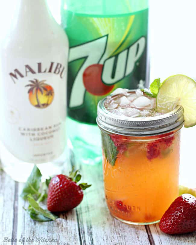A bottle of rum, 7UP, and a mason jar filled with strawberry mojito, fresh strawberries, mint, and lime slices on the rim