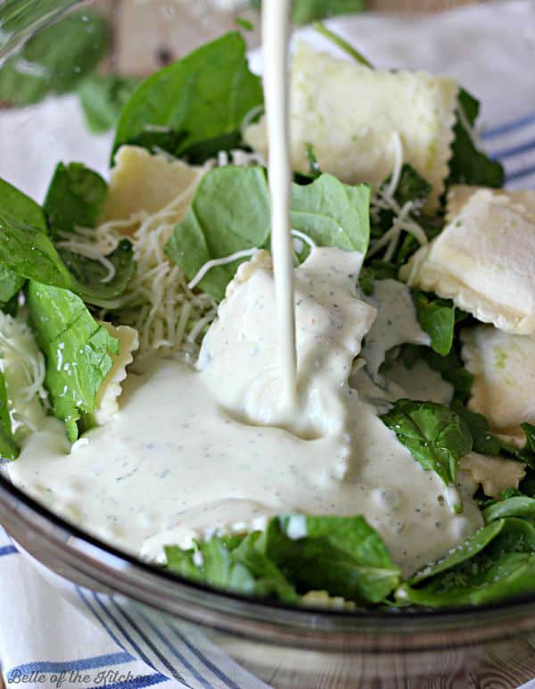 Alfredo sauce being poured over a bowl of spinach, ravioli, and shredded cheese