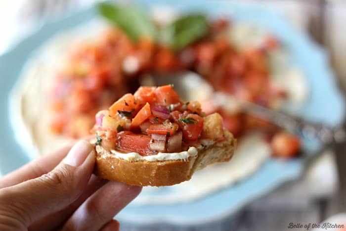 A hand holding a piece of crostini with cream cheese topped with bruschetta and basil