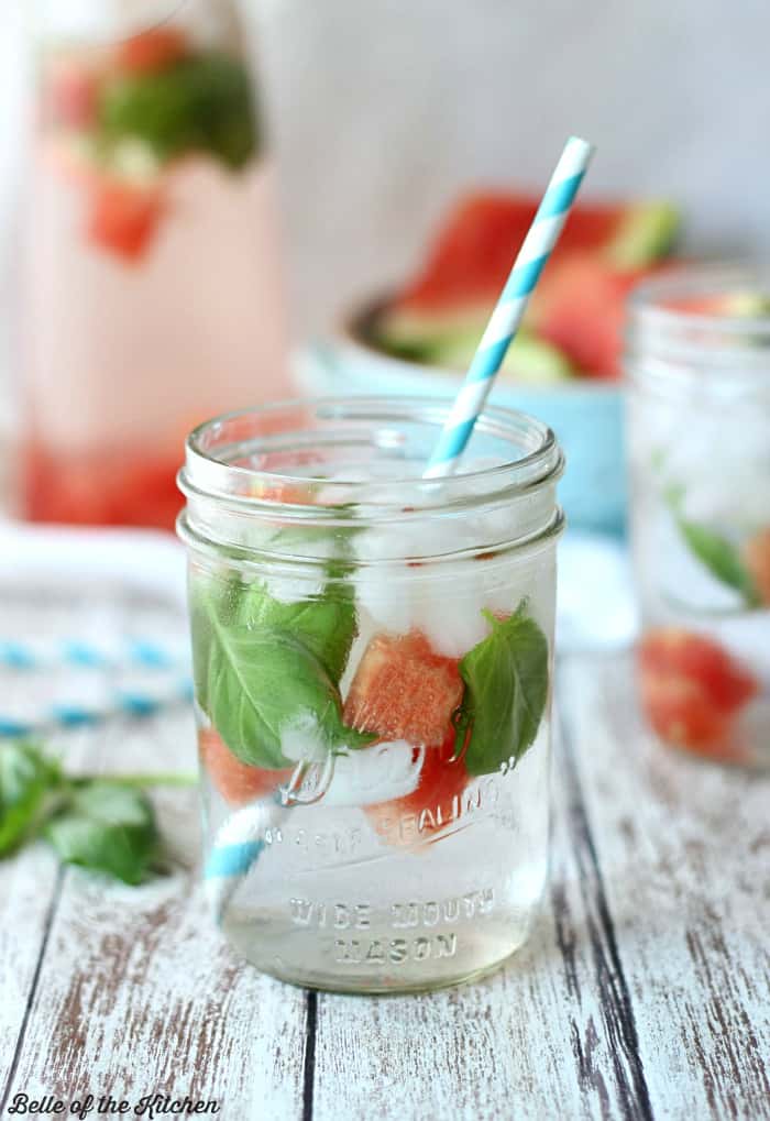 A close up of a glass cup on a table, with Basil, Watermelon, and water