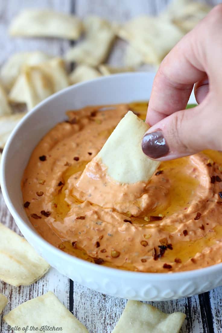 A bowl of red pepper dip surrounded by pita chips with a hand dipping in a chip