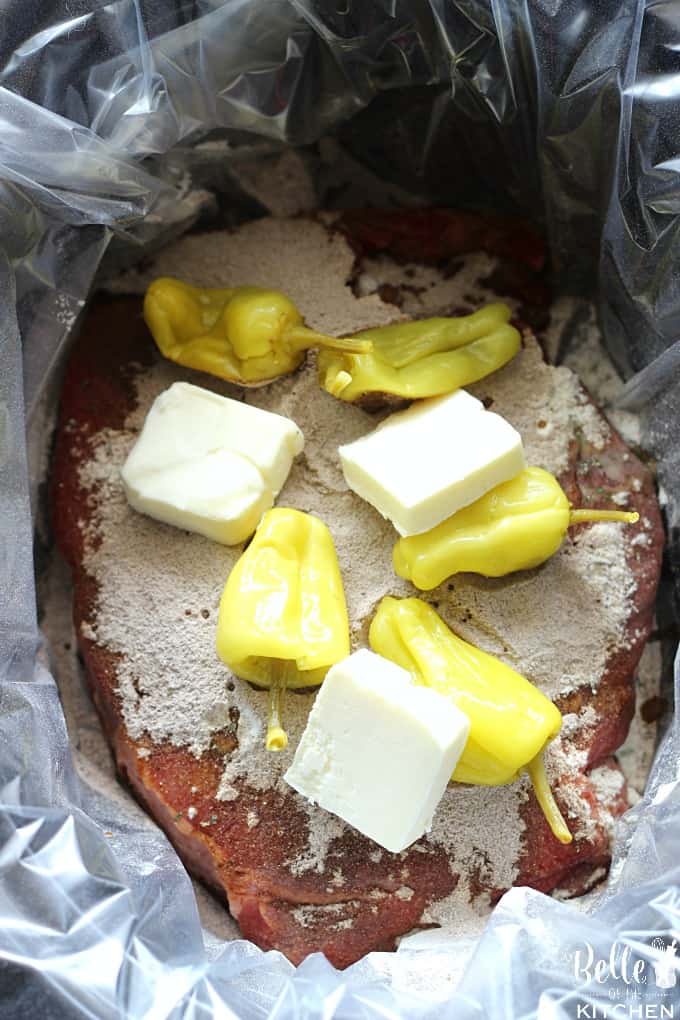 A crockpot filled with a chuck roast, seasoning, peperoncini peppers, and sliced butter