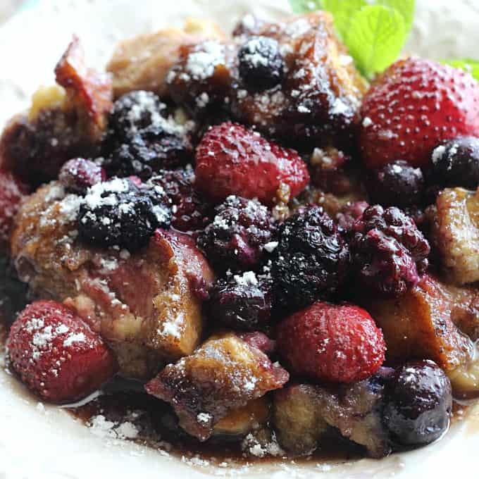 A plate of French toast casserole with berries on top and a sprig of mint on the side