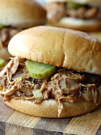 A close up of a sandwich with pulled chicken and pickles on a wooden cutting board