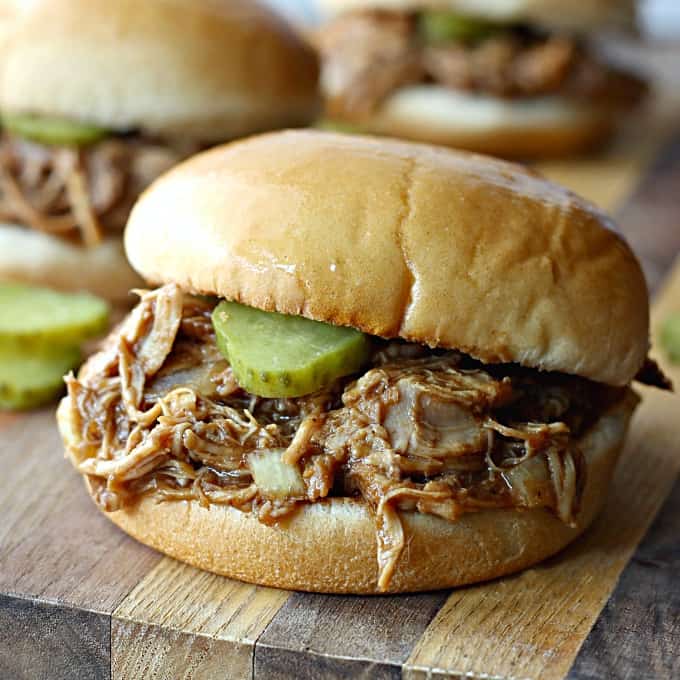 A close up of a sandwich with pulled chicken and pickles on a wooden cutting board