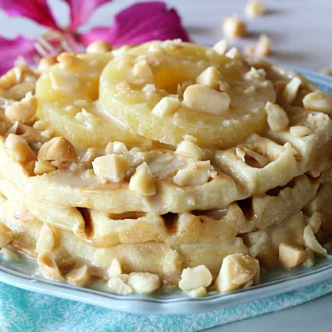 A close up of a plate of waffles with macadamia nuts and pineapple with a flower in the background