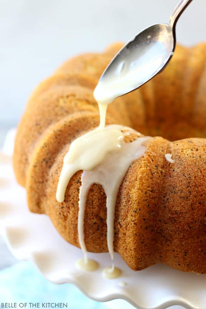 a lemon bundt cake with poppyseed vanilla glaze being drizzled on top with a spoon