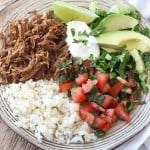 A plate of food with rice and vegetables, pork, lime, sour cream, and avocado