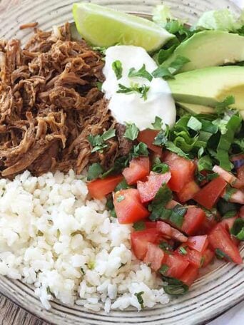 A plate of food with rice and vegetables, pork, lime, sour cream, and avocado