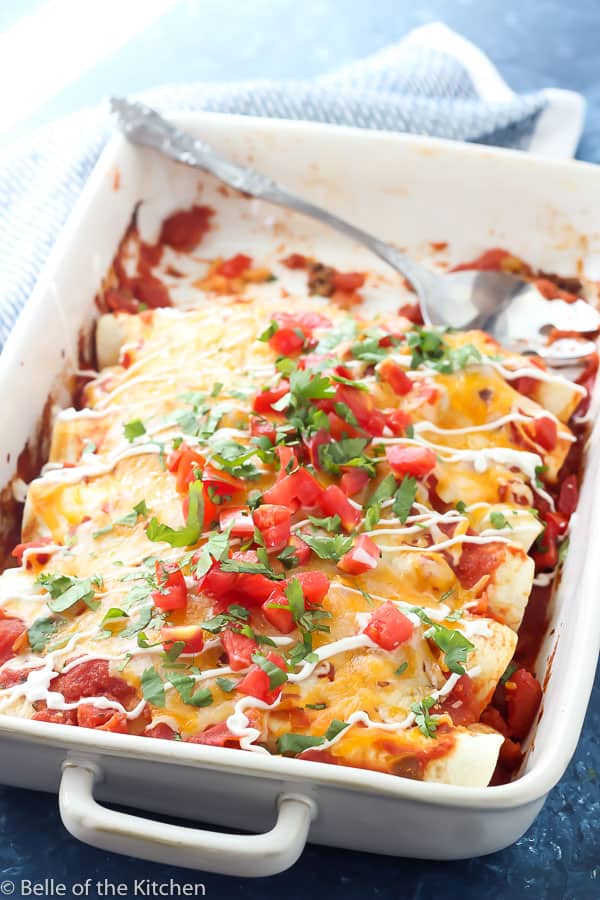 white rectangle casserole dish with enchiladas, diced tomatoes, cilantro, and sour cream with silver spoon