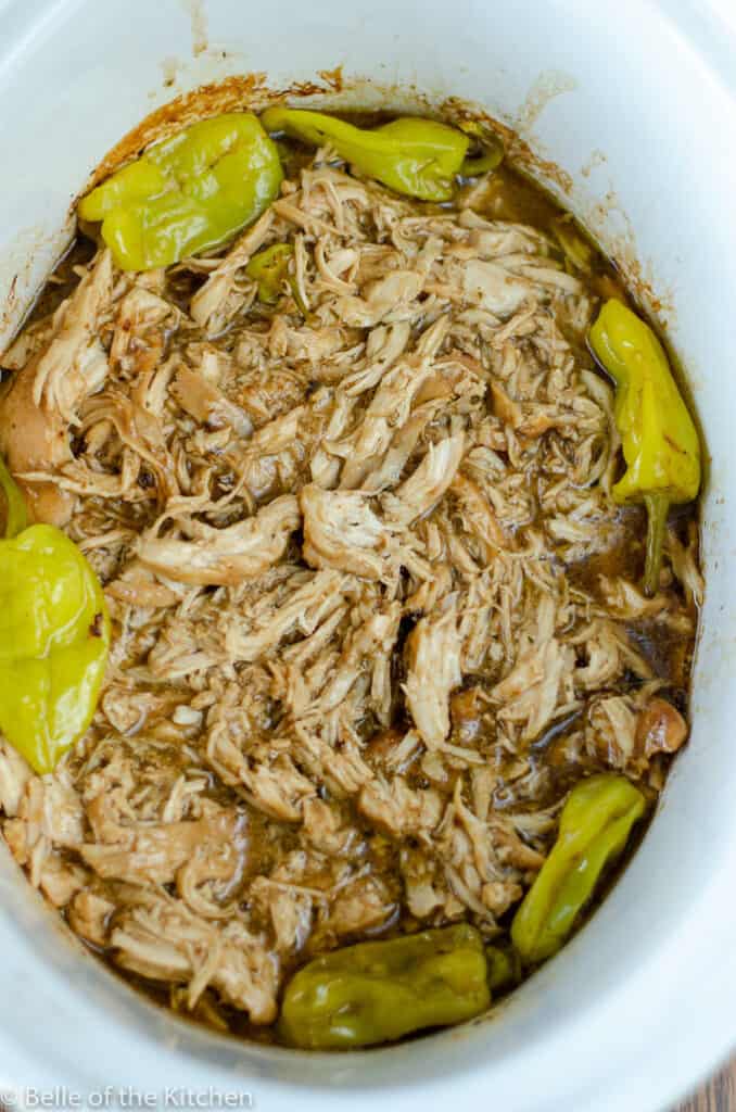 shredded chicken in a crockpot bowl surrounded by peperoncini peppers