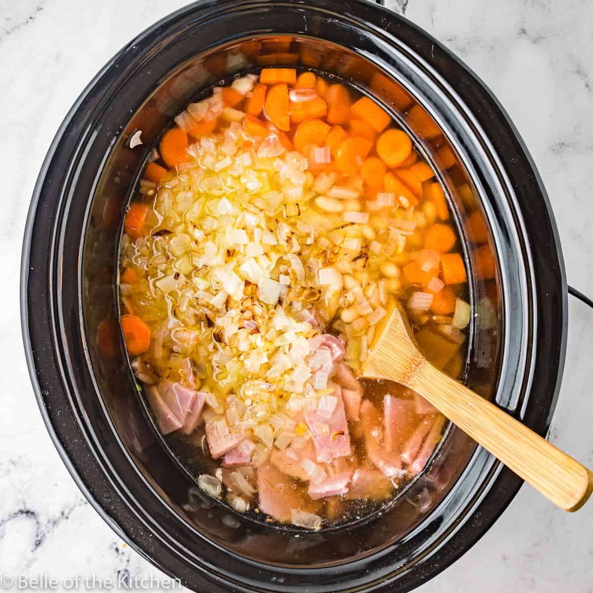 a crockpot filled with carrots, onions, beans, and ham, and a wooden spoon