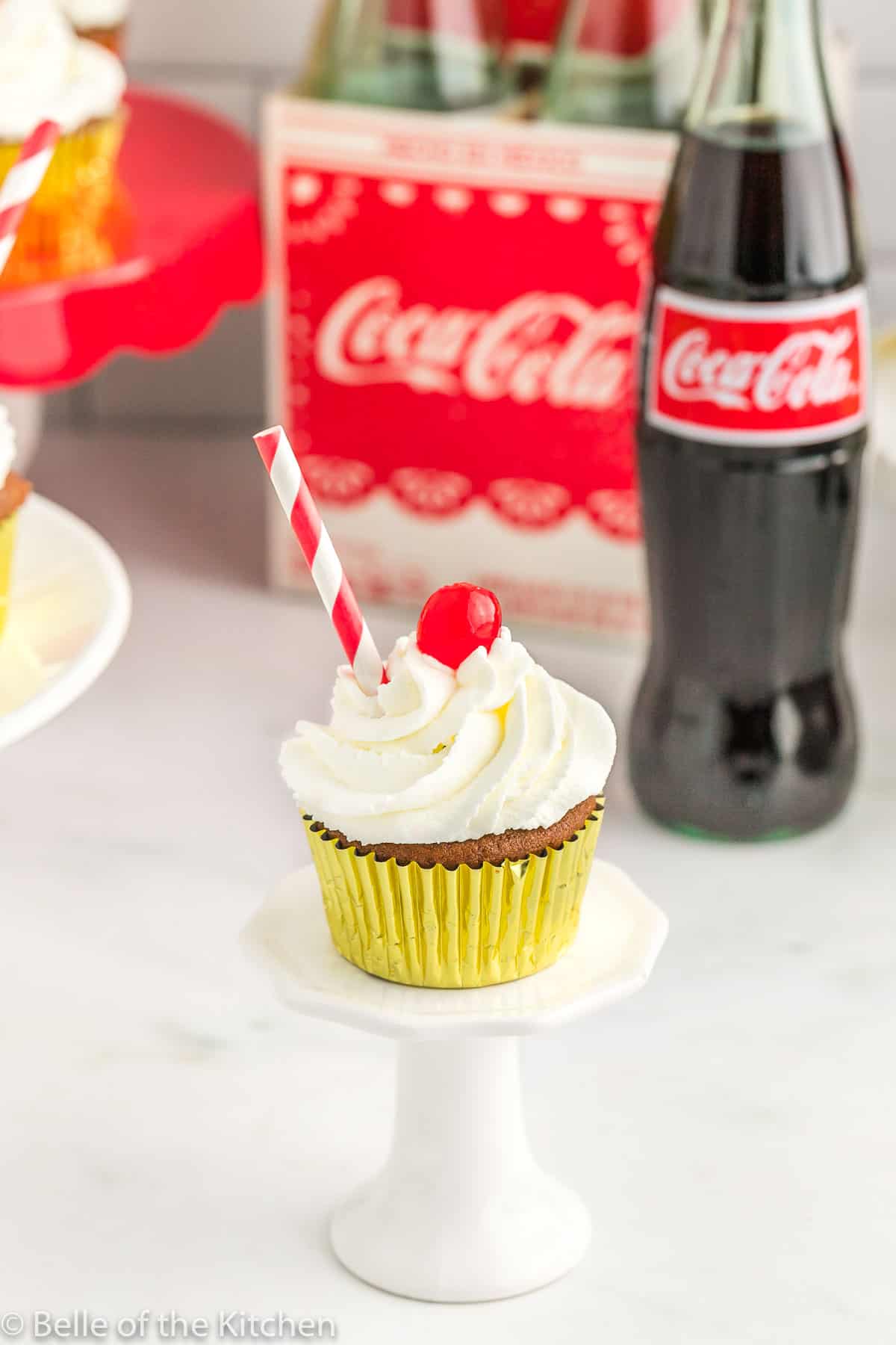 one cupcake on a cake stand next to a coke bottle