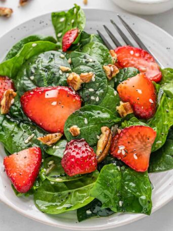 a white plate with a spinach salad topped with strawberries and a fork
