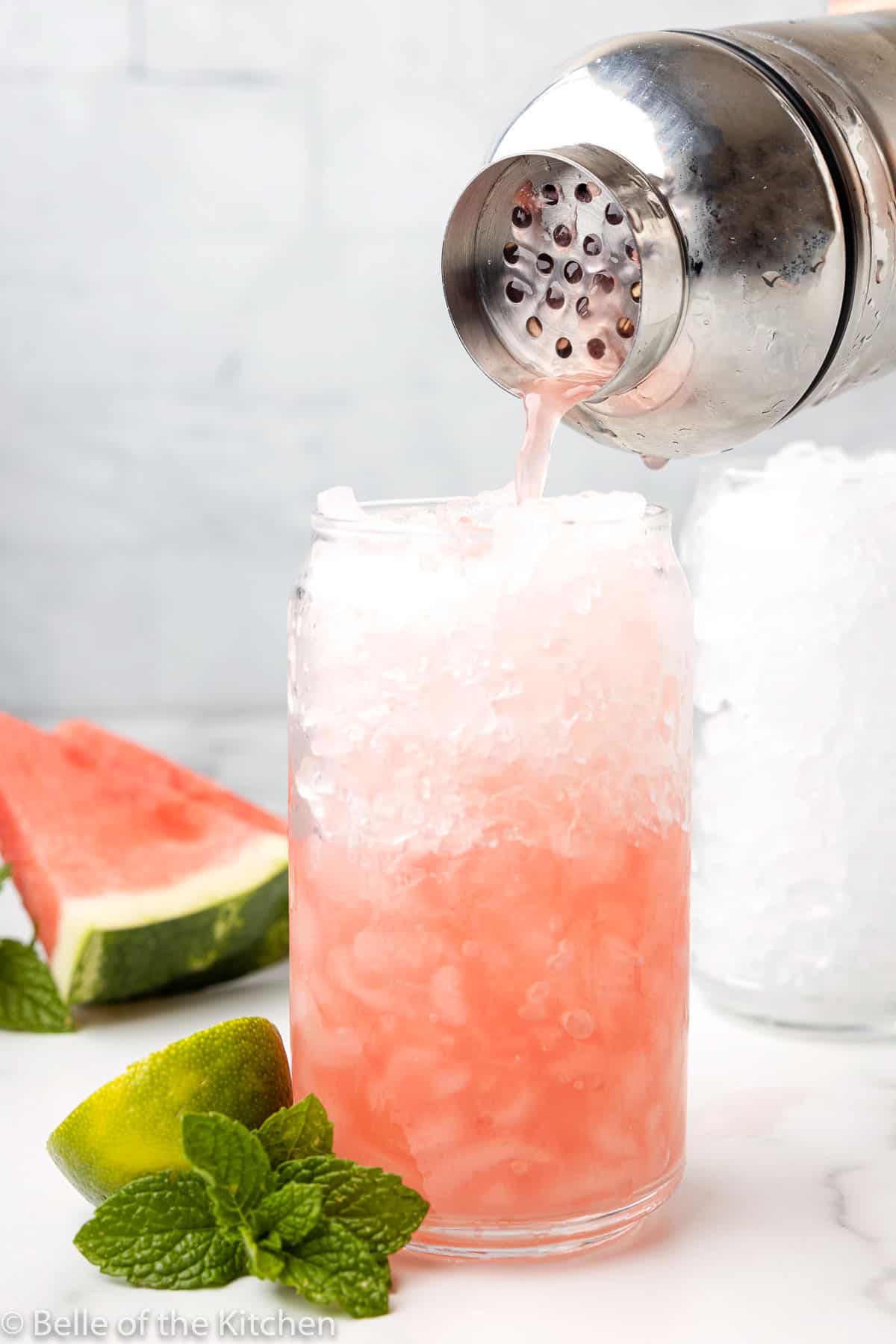 a cocktail shaker pouring into a glass of ice next to a slice of watermelon.