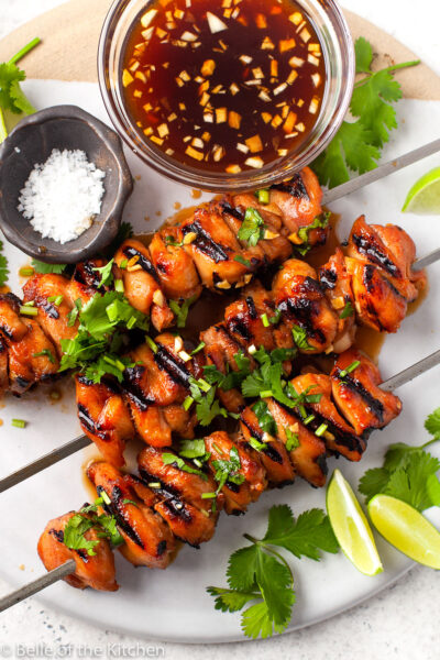 chicken skewers on a marble plate next to bowls of marinade and salt with limes on the side.