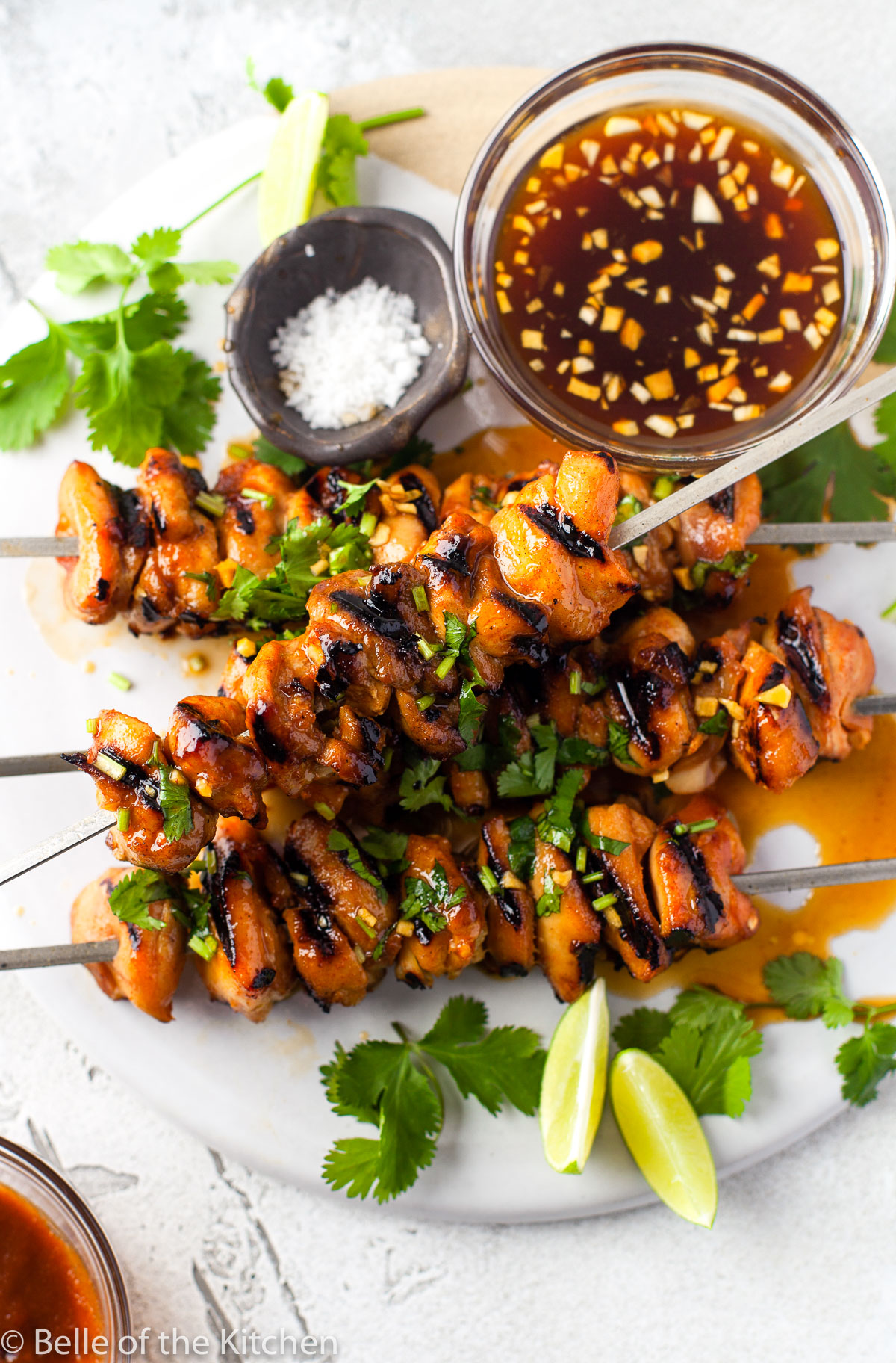chicken skewers on a marble plate next to bowls of marinade and salt with limes on the side.