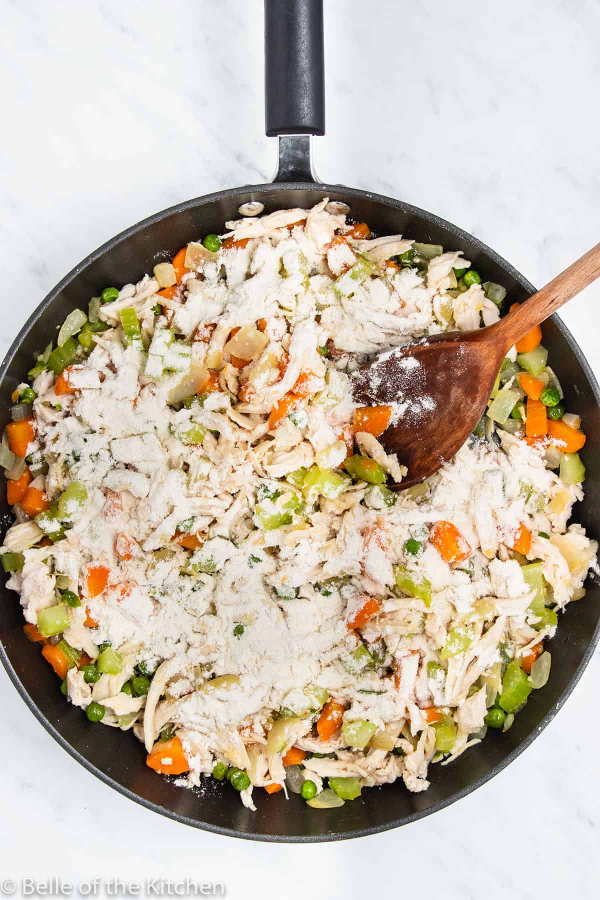 a skillet full of vegetables, chicken, flour, and a wooden spoon.
