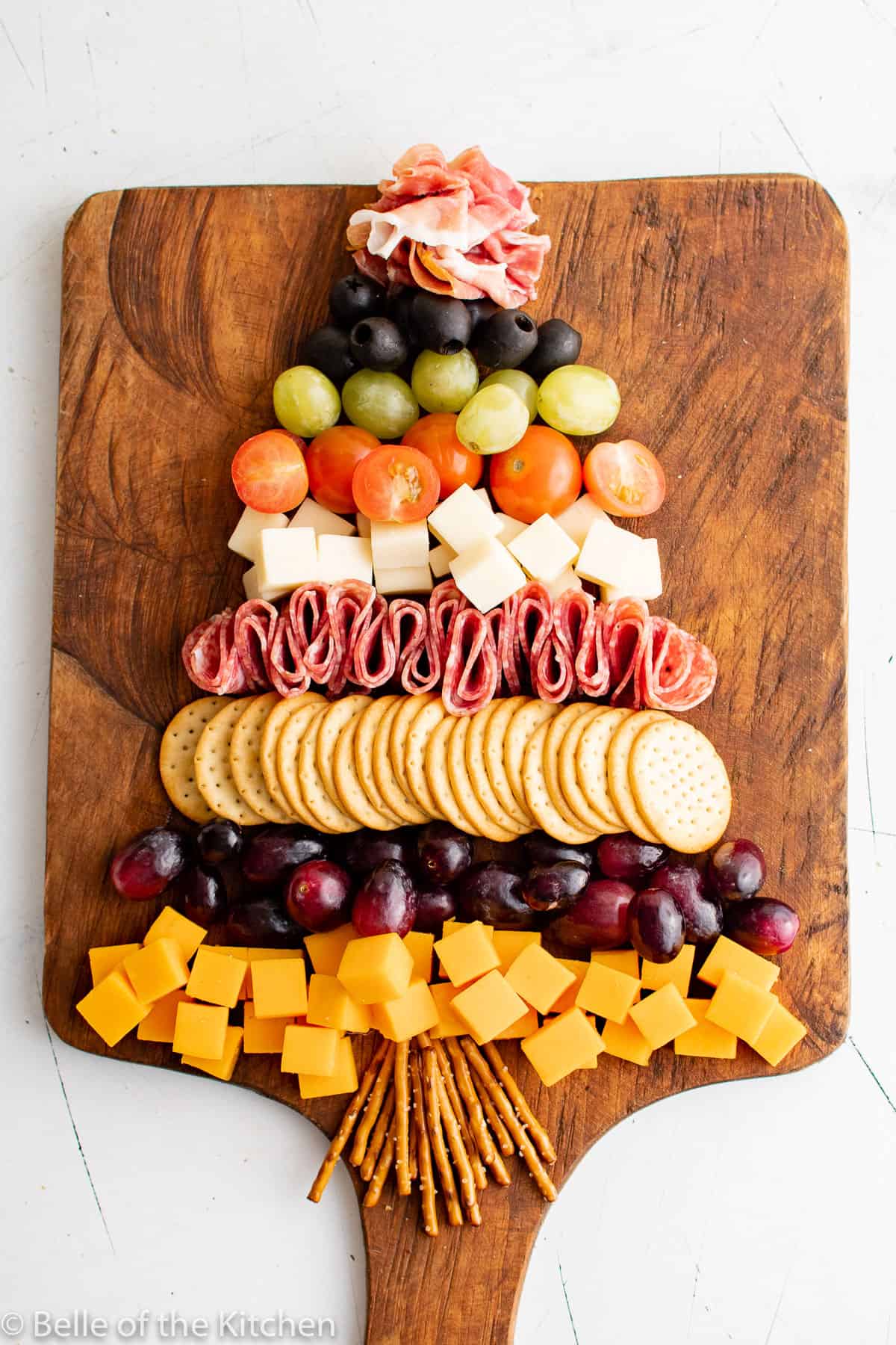 cut up cheese cubes, grapes, crackers, tomatoes, olives, prosciutto, and salami on a wooden cutting board.