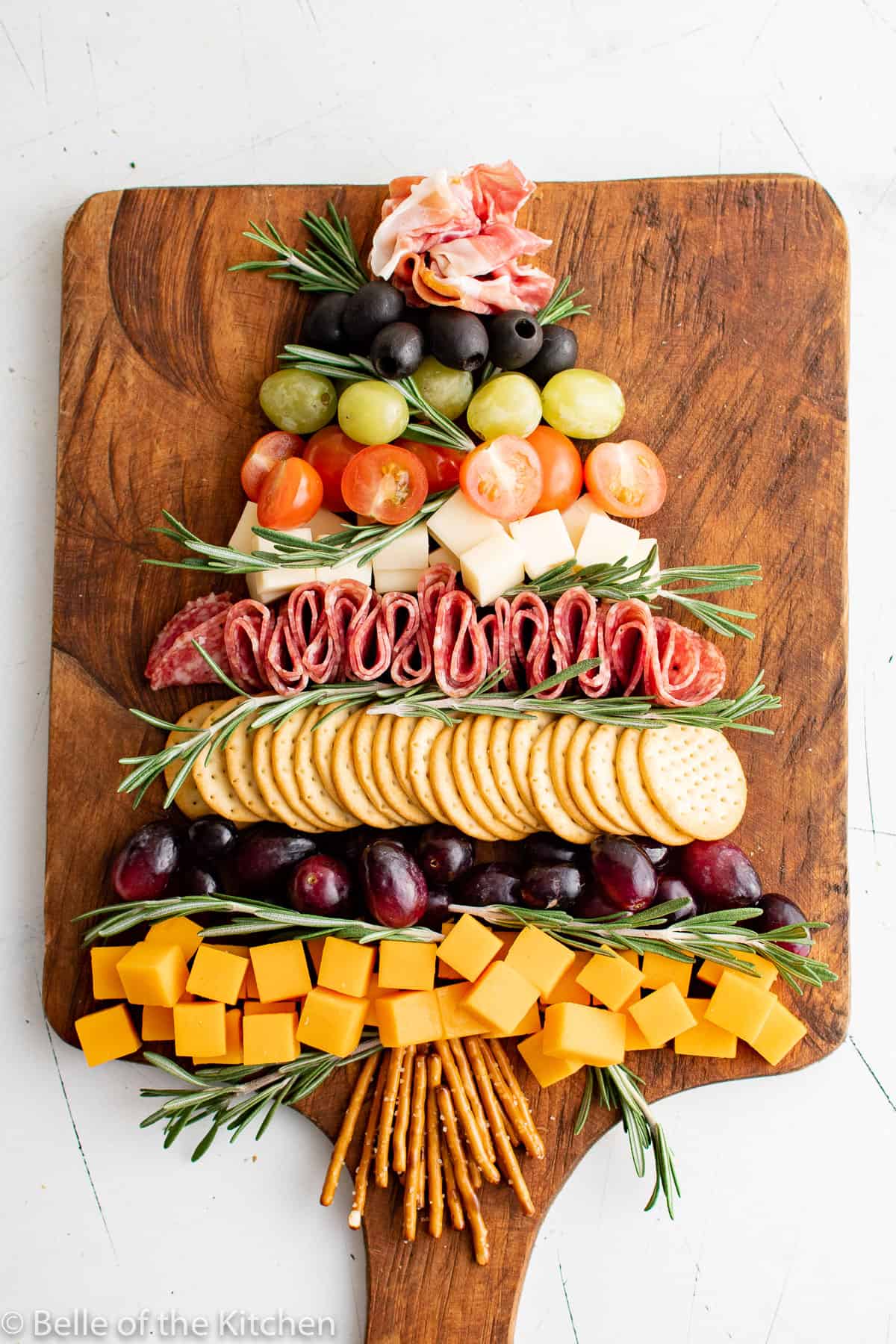 cut up cheese cubes, grapes, crackers, tomatoes, olives, prosciutto, and salami on a wooden cutting board in the shape of a Christmas tree.