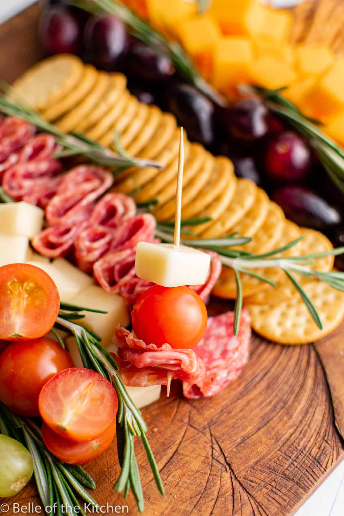 a toothpick holding a grape tomato and a cube of cheese next to a charcuterie board.