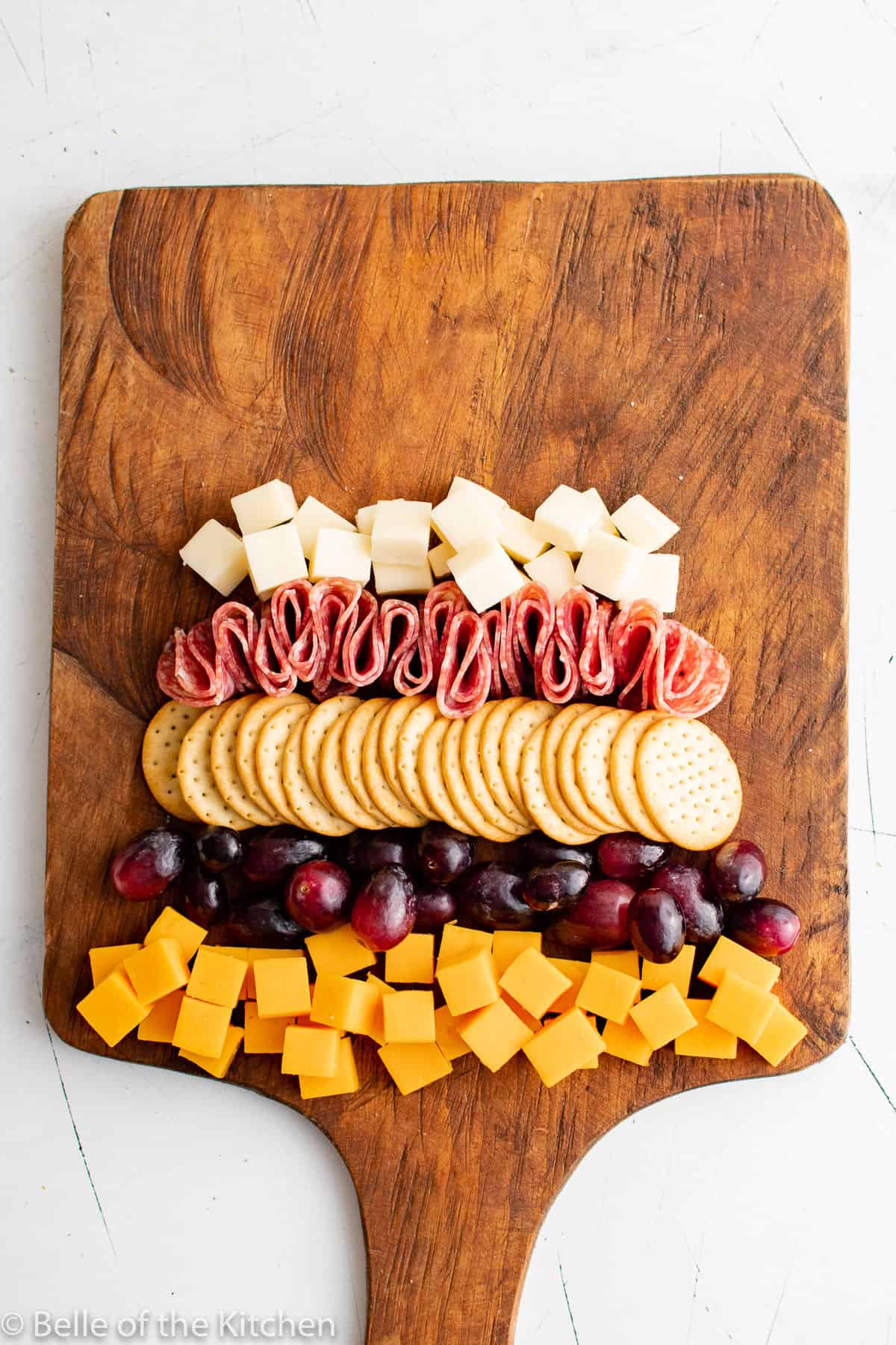 cut up cheese cubes, grapes, crackers, and salami on a wooden cutting board.