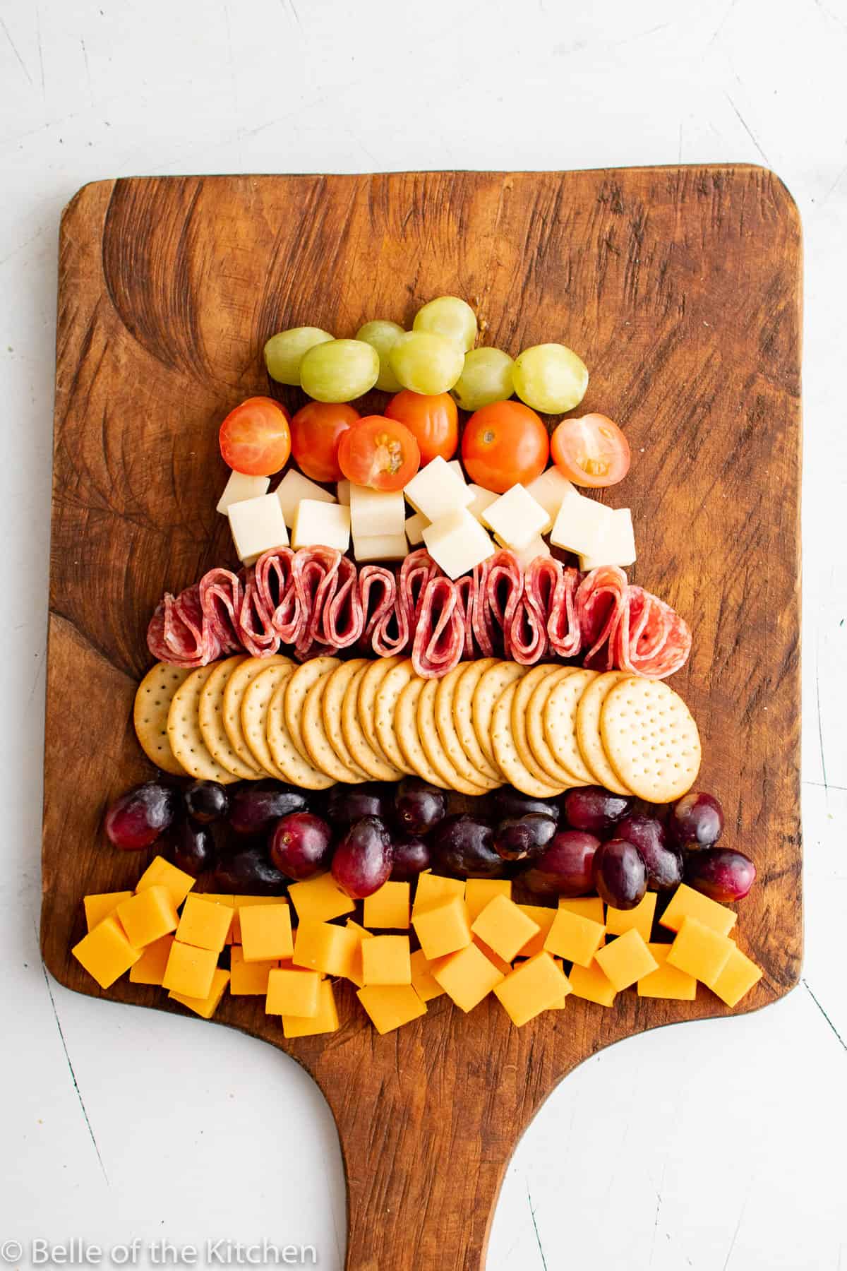 cut up cheese cubes, grapes, crackers, tomatoes, and salami on a wooden cutting board.