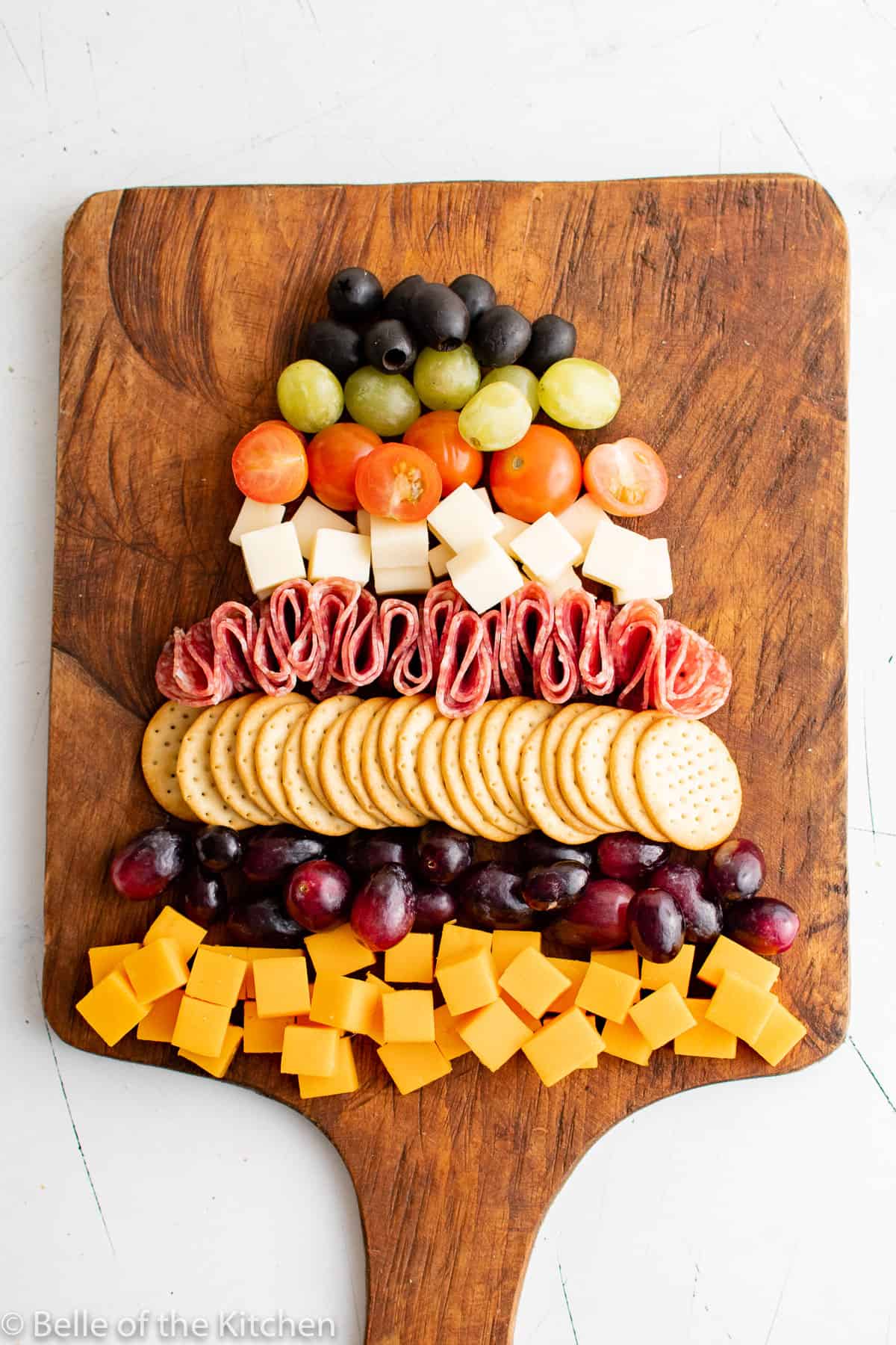 cut up cheese cubes, grapes, crackers, tomatoes, olives, and salami on a wooden cutting board.