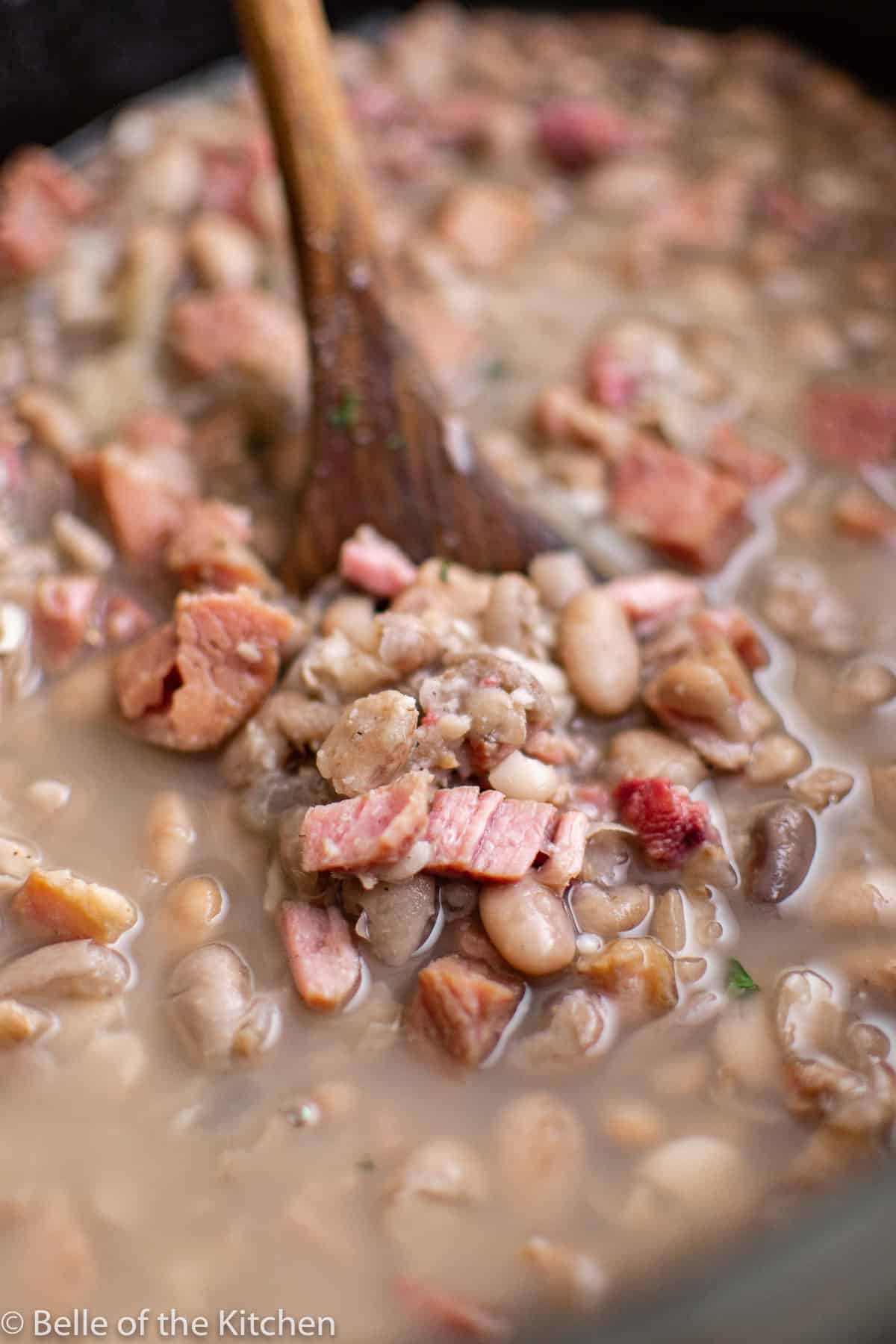 a crockpot bowl full of cooked ham and beans with a wooden spoon scooping some up.